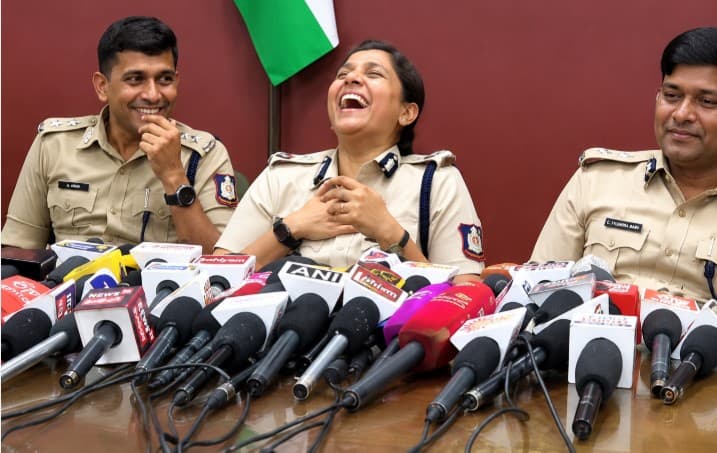 Indian police officers share a light-hearted moment during a press conference, seated behind multiple media microphones, with the national flag visible in the background.
